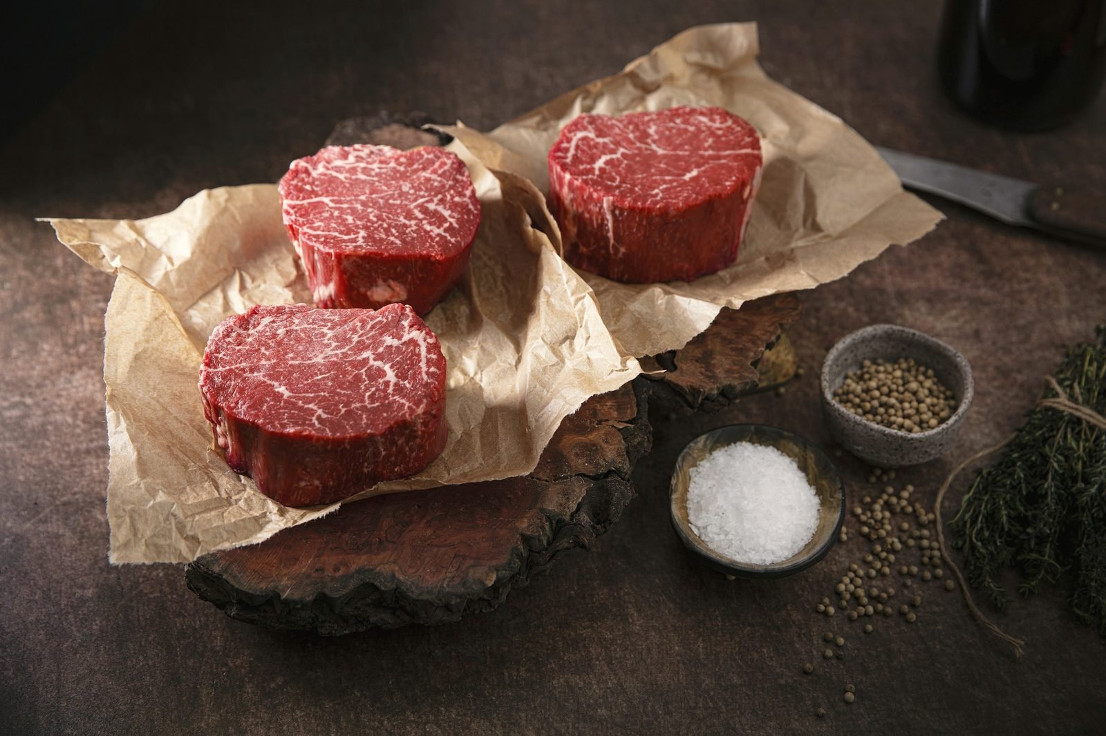 Three thick-cut beef filets sitting on crumpled brown paper with salt and peppercorns