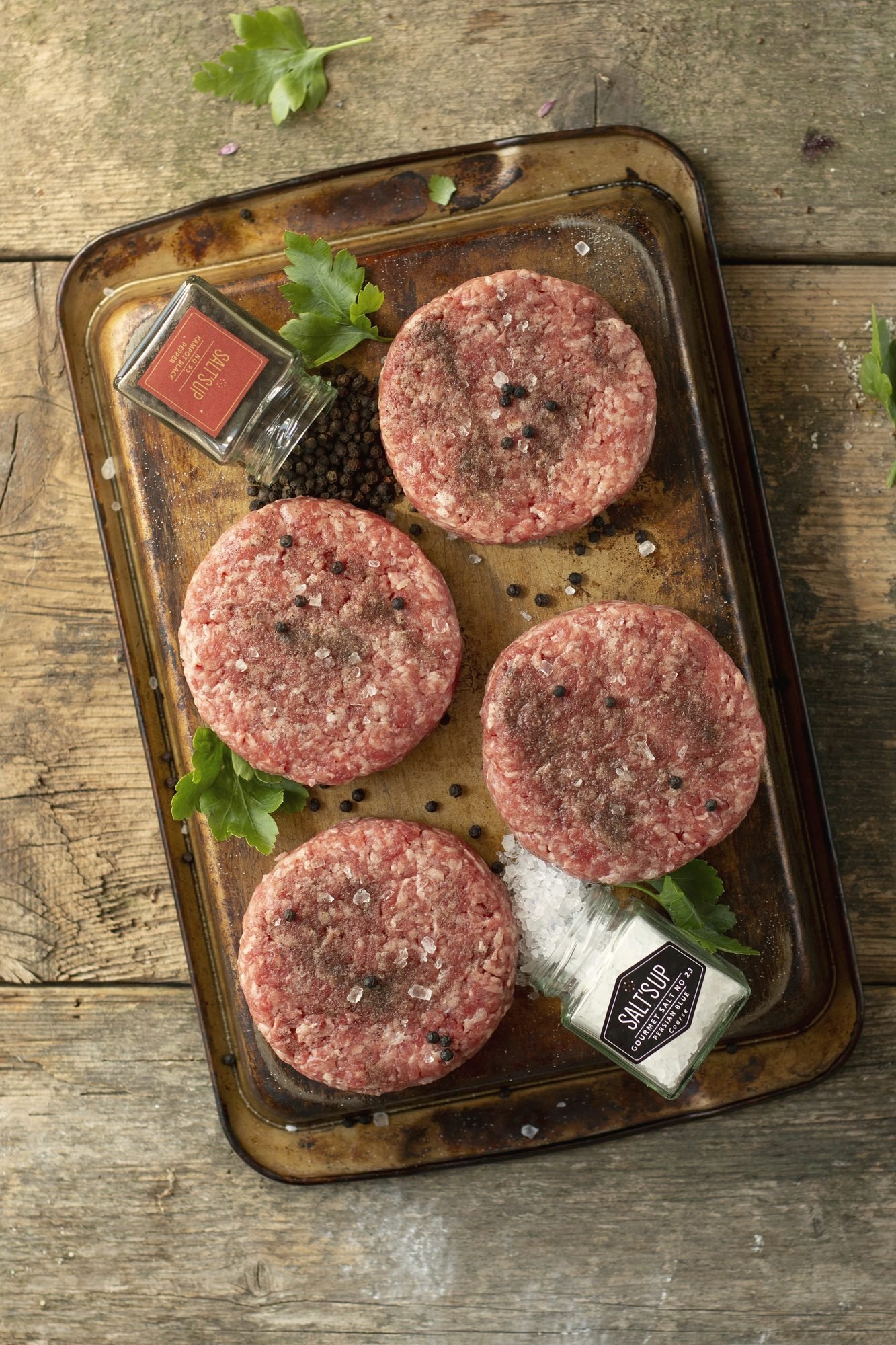 Four raw ground beef patties on a tray with herbs