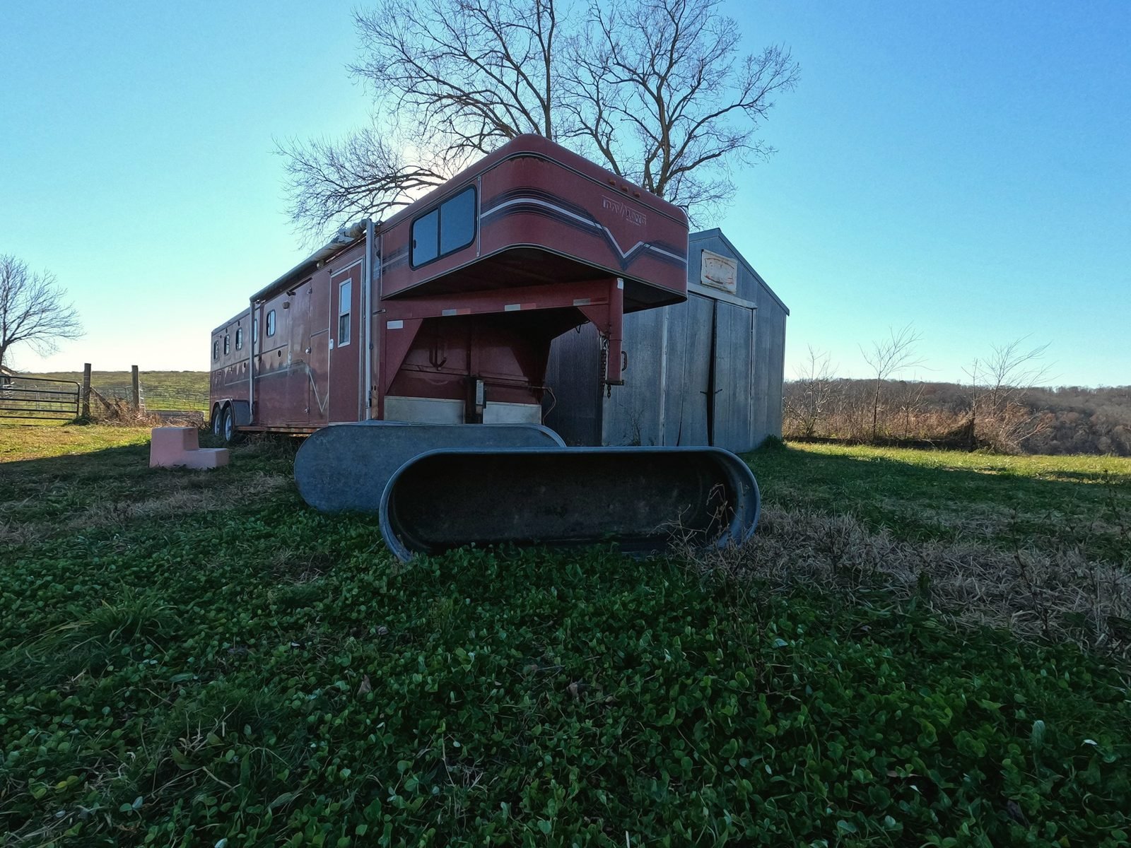 A red gooseneck livestock trailer parked on grass at a Missouri farm under a bare oak